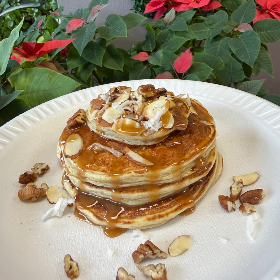 Stack of golden pancakes drizzled with caramel, topped with nuts and coconut flakes, against a plant background.