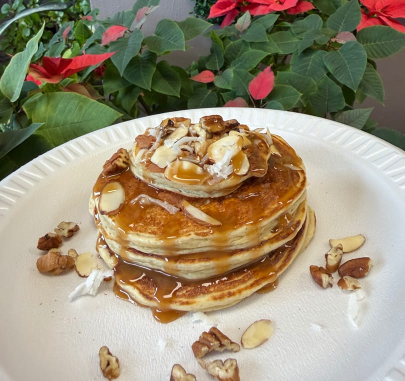 Stack of golden pancakes drizzled with caramel, topped with nuts and coconut flakes, against a plant background.
